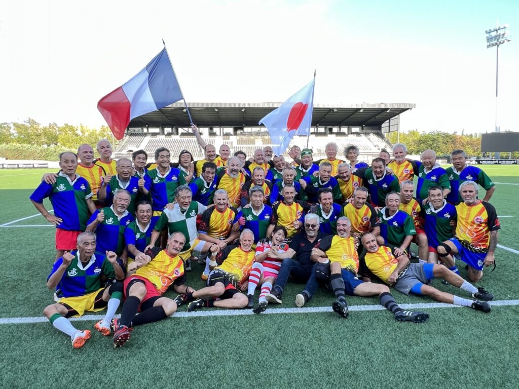 Photo de groupe des deux équipes de plus de 60 ans : Aix Baabaas et Kumamoto-Fukuoka mélangées après avoir joué l'une contre l'autre sur le terrain d'honneur du stade Maurice David, à Aix-en-Provence, France lors de la Coupe du Monde de Rugby 2023. Un drapeau japonais et un drapeau français sont tenus au dessus du groupe. Les joueurs japonais portent un maillot à gros damier bleu et vert, les français un maillot aux couleurs des deux clubs constitutifs des Baabaas (Esquiche-Coudes du Pays d'Aix et Tomtom Club) jaune, rouge et noir. Les joueurs du fond sont debout, ceux au milieu ont un genou à terre, ceux au premier rang sont assis ou allongés.