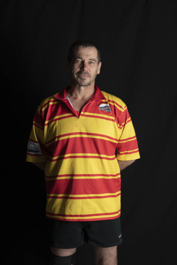 Portait de Christophe Raynaud-Leriche. Il porte le maillot de match des Esquiche-Coudes, aux couleurs de la Provence rayé jaune et rouge, un short noir, des chaussures de rugby rouges, une chaussette rouge à la jambe gauche et une chaussette noire à la jambe droite en hommage aux Esquiches déjà disparus. La photo a été prise par Karine Joly en 2023 avant un match contre les Tomtom au Stade Maurice David à Aix-en-Provence.