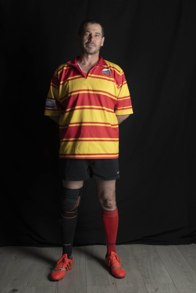 Portait en pied de Christophe Raynaud-Leriche. Il porte le maillot de match des Esquiche-Coudes, aux couleurs de la Provence rayé jaune et rouge, un short noir, des chaussures de rugby rouges, une chaussette rouge à la jambe gauche et une chaussette noire à la jambe droite en hommage aux Esquiches déjà disparus. La photo a été prise par Karine Joly en 2023 avant un match contre les Tomtom au Stade Maurice David à Aix-en-Provence.